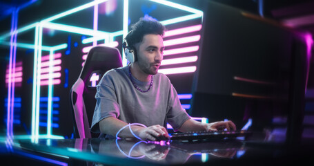 Young Man in a Futuristic Neon Room Playing Action Video Games on a Computer. Handsome Male Putting On Headphones and Talking to Online Stream Audience. Streaming and Gaming Concept © Gorodenkoff
