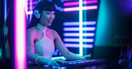 Portrait of a Happy Asian Girl in Headphones Talking with Friends Online on a Computer. Smiling Streamer or Video Gamer Playing Games and Chatting with Internet Followers in a Futuristic Neon Studio