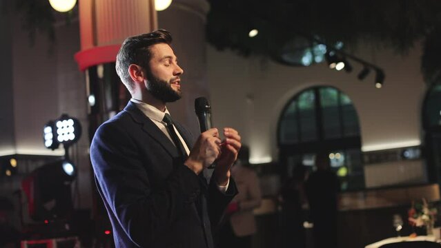 Young caucasian wedding host dressed in black suit taking word and speaking in front of audience in fancy restaurant. Bearded man with microphone in hands expressing best wishes to newlyweds.