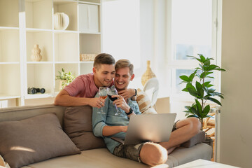 Young gay couple sitting in an apartment on a sofa using a laptop and drinking wine. Modern gay family concept