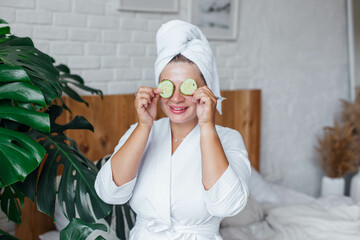 A young beautiful smiling woman in a white robe and a towel on her head holds cucumber slices...
