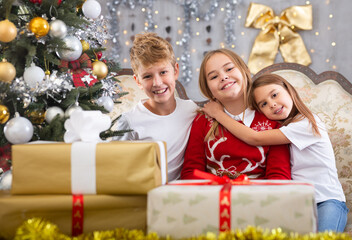 Fototapeta na wymiar Happy brother with two sisters are sitting on sofa together with gift boxes next to the new year tree