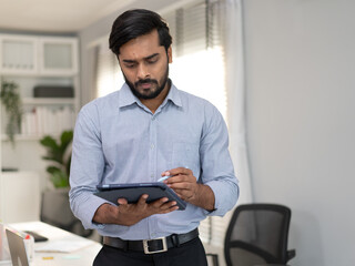 Success Indian businessman standing in modern office writing note on tablet. Confident professional...