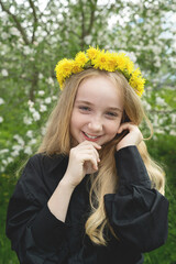 Fototapeta premium A girl of 13 with a wreath of dandelions in a spring flowering garden. The girl smiles and looks at the camera