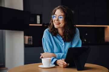 Smiling woman drinking coffee and using tablet computer while sitting in kitchen