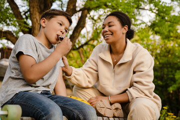 Fototapeta premium Little boy eating candy bar while sitting with his mother in park