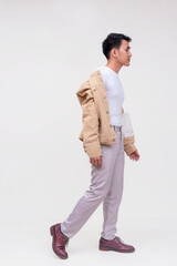 Fototapeta premium A casual FIlipino guy in a white shirt and light gray pants and a half worn khaki jacket takes a calm relaxed stroll. Whole body photo, isolated on a white background.