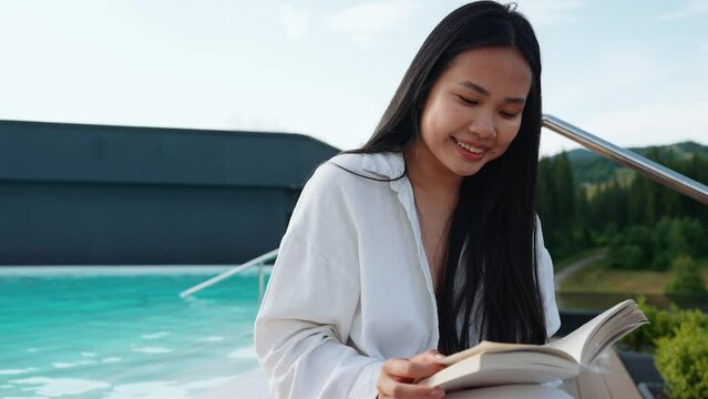 Cheerful asian woman relaxing and reading book by pool at mountain resort on balcony in summertime