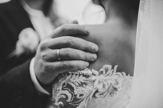 Groom gently embraces bride by shoulders closeup. Newlyweds together hugging in nature. Back view. Man hugs woman stand on wedding ceremony. Couple in country. Details of moment. Black and white photo