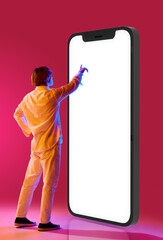 Young guy standing next to giant 3d model of smartphone and tapping on screen against pink studio background in neon lights. Concept of human emotions, facial expression, youth, lifestyle. Ad
