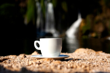 White mug standing on rocky beach next to the waterfall. Beautiful nature destination. Coffee or...