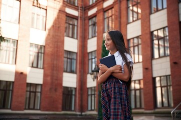 Standing against the building. School girl in uniform is outdoors