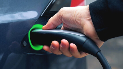 close-up view of hand holding an electric vehicle charging gun connector and plugging it into charger port of electric car. EV charge process parts concept background.