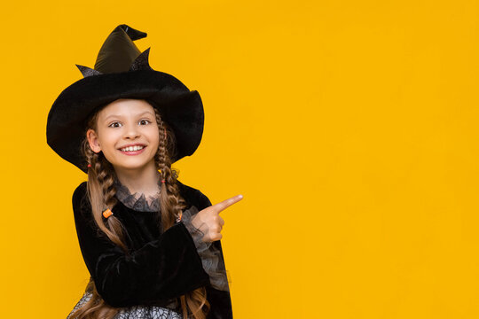 A surprised girl in a witch's fancy dress and with long pigtails points to the side at an advertisement, a place for advertising with a cute witch, a witch on a yellow isolated background.