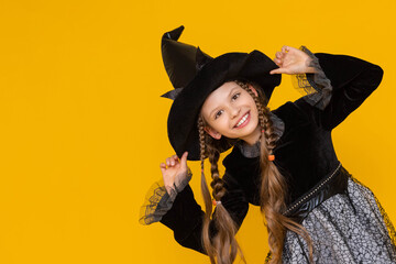 A young witch in a black fluffy dress and long pigtails supports her pointed black hat with her...