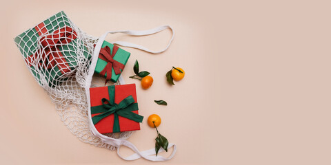 Gifts with ribbons are in a white eco-friendly string bag on a beige background, next to orange ripe tangerines with leaves. Top view. Copy space