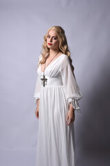 Fototapeta premium Close up portrait of beautiful blonde model wearing elegant white halloween gown, a historical fantasy character. Holding crucifix cross necklace, isolated on studio background.