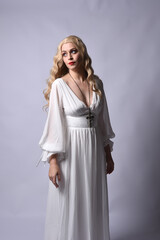 Fototapeta premium Close up portrait of beautiful blonde model wearing elegant white halloween gown, a historical fantasy character. Holding crucifix cross necklace, isolated on studio background.