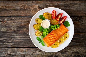 Seared salmon steak with baked potatoes and fresh vegetable salad served on wooden table
