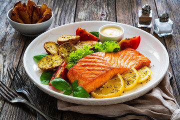 Seared salmon steak with baked potatoes and fresh vegetable salad served on wooden table
