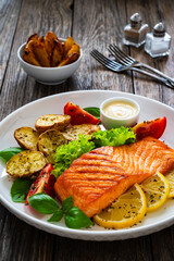 Seared salmon steak with baked potatoes and fresh vegetable salad served on wooden table
