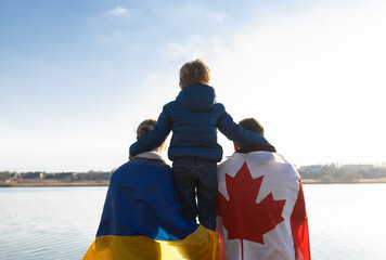 woman with Ukrainian flag, man with Canadian flag behind back and child , sitting with backs...