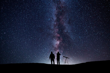 Fantasy landscape, two hiker standing on the hill, and looking at the Milky Way galaxy with telescope.