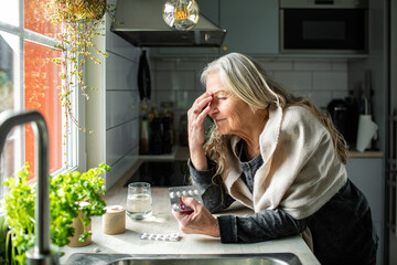 Senior woman taking medication for a migraine in the kitchen at home