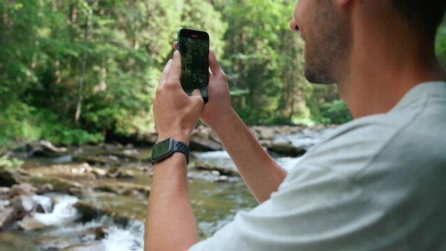 Back view of happy man records video of mountain river on cell phone in forest 