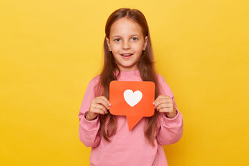 Popular child content, blogging. Portrait of charming preschool girl holding heart like icon of social media and looking at camera with toothy smile isolated over yellow background