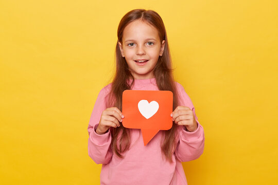Popular child content, blogging. Portrait of charming preschool girl holding heart like icon of social media and looking at camera with toothy smile isolated over yellow background