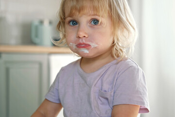 Baby girl enjoying ice cream. Pretty little toddler eating an ice-cream indoors, at home. Dining room background. Small child eats plombir and cream messy on her mouth. Cute kid with tasty sweet food.