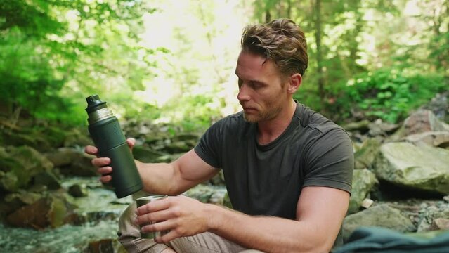 Handsome blond tourist man drinking tea from thermos near mountain river in woods