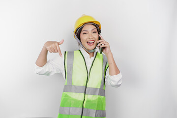 A smiling Asian woman labor wearing safety helmet and vest, talking on her phone and pointing to copy space below her, isolated by white background. Labor's day concept.