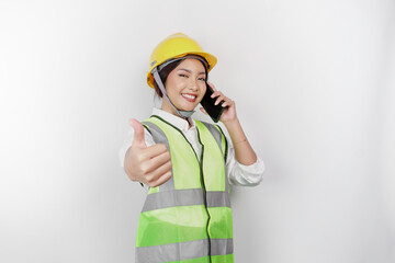 Excited Asian woman labor wearing safety helmet and vest gives thumbs up hand gesture of approval while having a phone call, isolated white background.