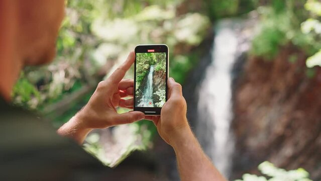 Back view of tourist man takes picture of waterfall on mobile in mountains