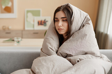 Portrait of depressed young woman wrapped in blankets at home struggling with mental health issues,...