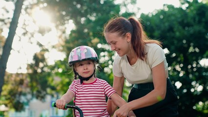 Mother teaches kid daughter to ride bike in park. Happy family, little girl in helmet together with her mother learns to ride bicycle outdoors in summer. Family weekend. Child cyclist with mom, nature