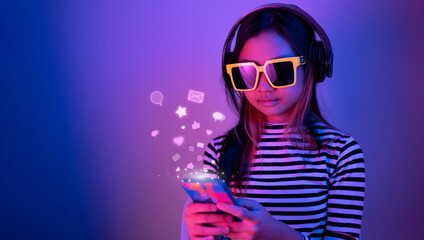 Young Asian teen wearing sunglasses and headphones using smartphone communicating with friends on social media in a room with blue and red neon lights. - obrazy, fototapety, plakaty