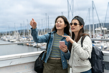 travel lifestyle and people in usa. two asian women looking at distance with pointing gesture while consulting information on phone near fishing port at Old Fisherman's Wharf in California