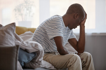 Side view portrait of adult Black man sitting on bed in morning struggling with depression, copy space