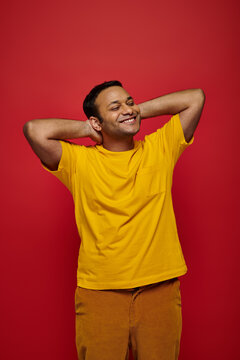 pleased indian man in bright casual clothes standing and smiling on red background, relaxed pose