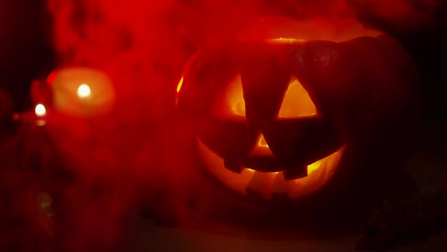 Handmade pumpkin jack o lantern lights up next to candles at night with sinister smile. Eerily flickering illumination of homemade Halloween decor appears from darkness in fog.