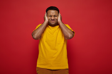 tensed indian man in bright clothes standing with closed eyes and covering ears on red background
