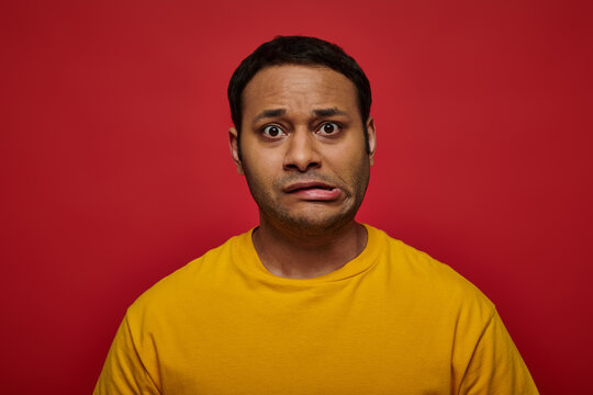 worried indian man in bright clothes looking at camera and grimacing on red background, stroke