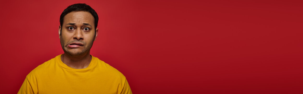 worried indian man in bright clothes looking at camera and grimacing on red background, banner