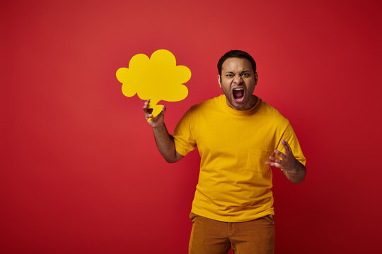 angry indian man in yellow t-shirt holding blank speech bubble and screaming on red background