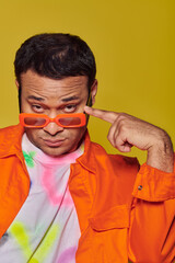 self-expression, confident indian man adjusting orange sunglasses on yellow backdrop, style icon