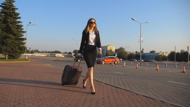 Business woman with suitcase walks to the airport to go on business trip. Lady in high heels shoes stepping with her luggage along street at sunset time. Travel concept. Slow motion Close up