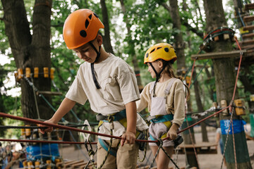 A little smiling girl and boy climbs a rope harness in a summer  rope park pass obstacles